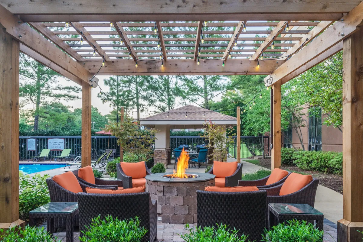 The image features an outdoor fire pit area with cozy seating, a pergola for shade, and landscaping details. The swimming pool is visible in the background, and the atmosphere is welcoming and set for relaxed gatherings, especially in the evening.