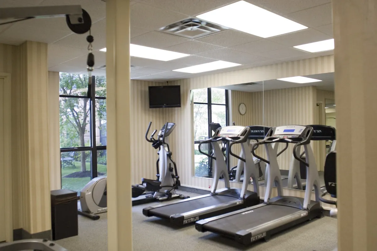 A bright, modern fitness center featuring several treadmills and an elliptical machine lined up by expansive windows. The space is filled with natural light and offers views of the outdoors, creating a motivating environment for exercise. There are also TVs mounted on the walls for entertainment during workouts.