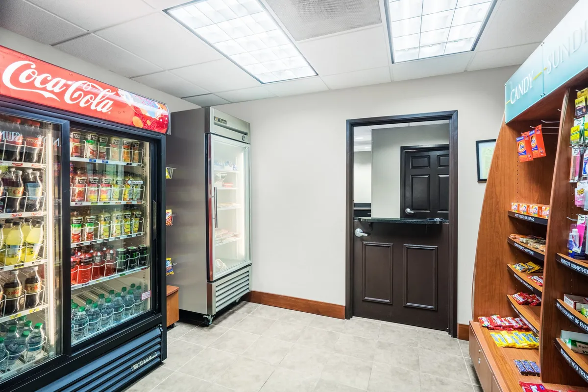 This image shows an inviting snack and beverage area featuring a Coca-Cola vending machine and a well-stocked snack display rack. The space is relaxed and casual, with a doorway visible that suggests easy access to adjoining amenities. The ambient lighting and organized setup make it appealing for quick, self-serve refreshments.