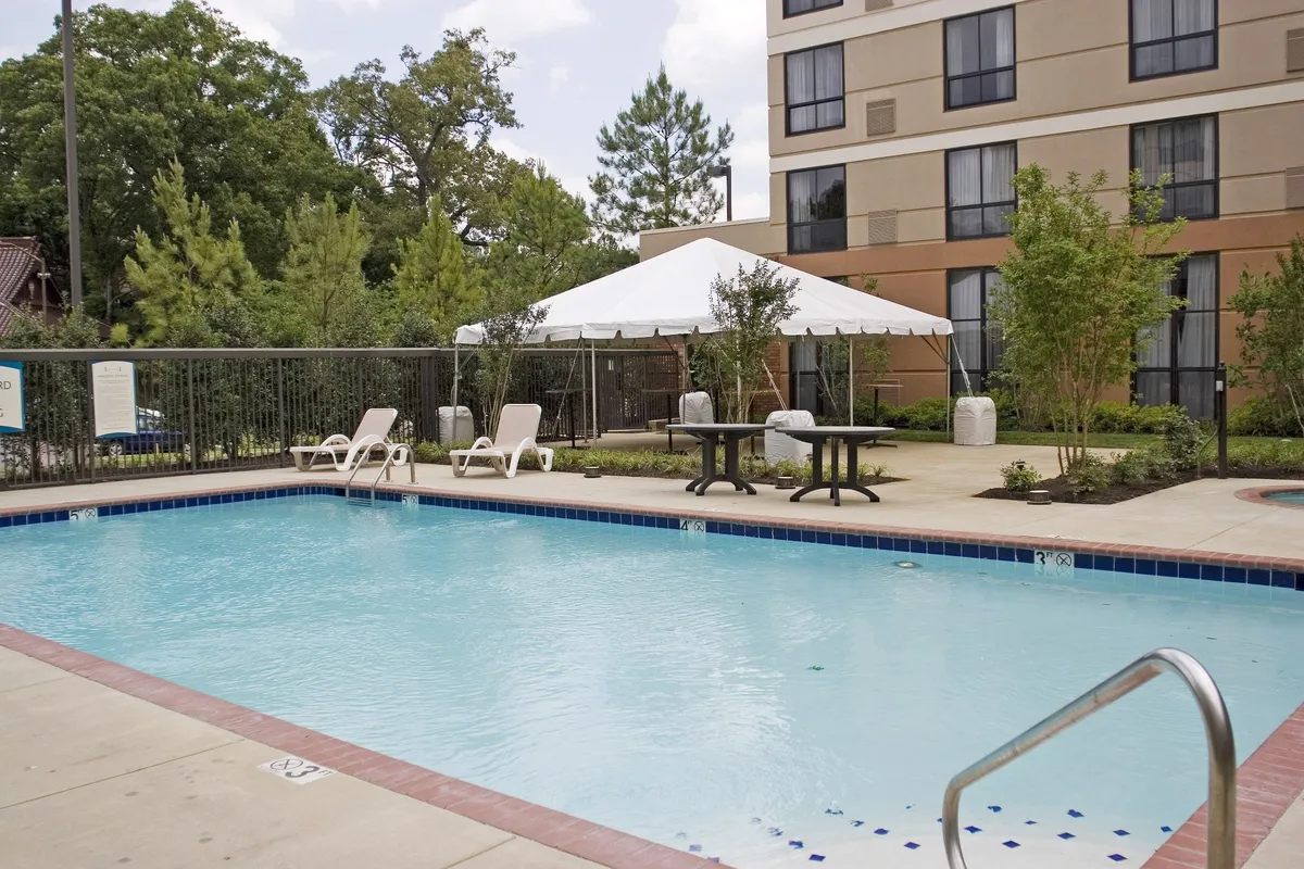 The image offers a different angle of the swimming pool area, featuring several lounge chairs, a tented space for shade, and well-maintained landscaped greenery. There are people enjoying the pool, gathering by the tent area, and soaking up the sun, evoking a vibrant, communal, and social mood.