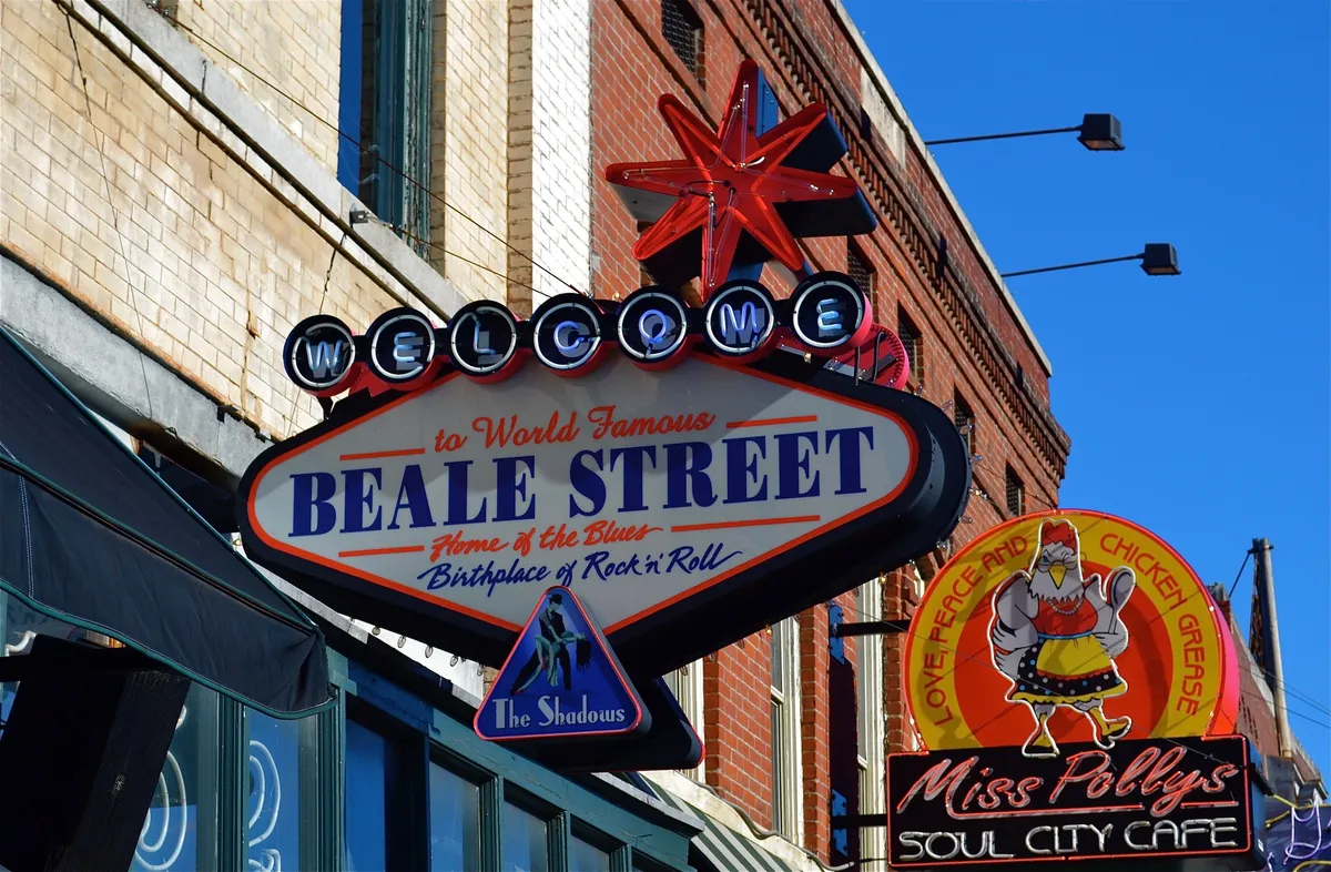 This lively exterior shot captures the vibrant entrance to Beale Street, featuring a prominent welcome sign, a Beale Street sign, and the colorful storefront of Miss Polly's Soul City Cafe. Visitors are seen enjoying the festive atmosphere, dining at local cafes, taking in the sights, and soaking up the unique Memphis vibe. The scene is bustling with energy, hinting at the many family-friendly attractions and experiences just steps away.