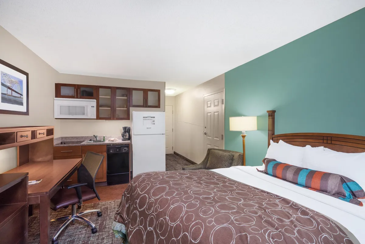 The image showcases a stylish studio suite with a plush queen bed, a convenient kitchenette for easy meals, and a bright workspace with a desk. The space feels inviting and practical, perfect for relaxing, catching up on work, or preparing a quick snack after exploring Memphis.
